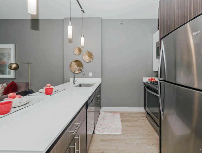 Modern kitchen with stainless steel appliances, a long white countertop with red accents, and wooden flooring. Two hanging lights illuminate the space. Wall art decorates the gray background.