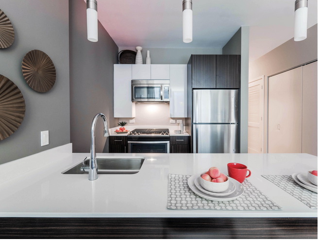 Modern kitchen with a white countertop, stainless steel appliances, and decorative plates on a gray wall. A red mug and plates with peaches are on the island.