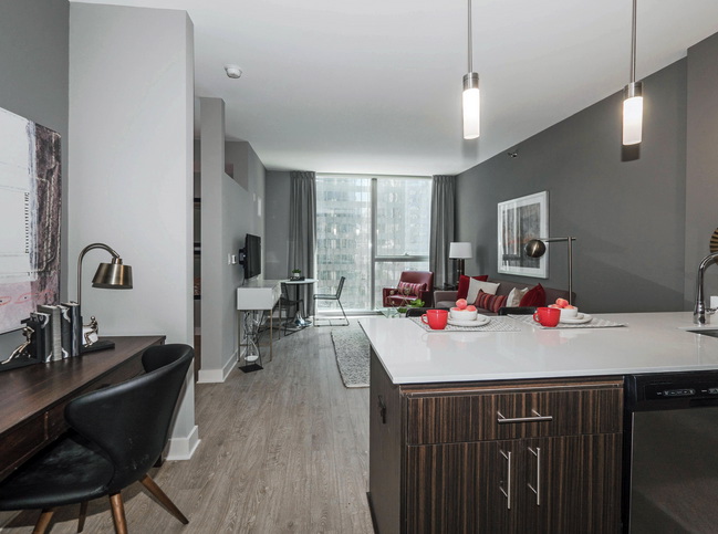 Modern apartment interior with an open-plan layout. Features a kitchen, white island, red seating area, desk, and large window. Neutral tones and contemporary decor throughout.
