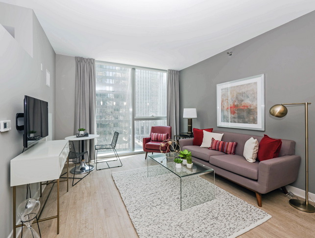 Modern living room with a gray sofa, glass coffee table, red armchair, and abstract art on the wall. A flat-screen TV and desk with chairs are near the window with city views.