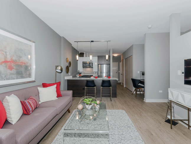 Modern open-concept living room and kitchen with gray walls, a sofa with red accents, glass coffee table, and bar stools by a countertop.