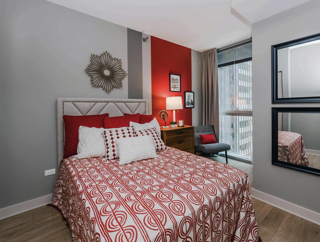 Bedroom with a red and white patterned bedspread, decorative pillows, and a red accent wall. Features a sunburst wall decor, bedside lamp, light-filled window, and two black-framed mirrors.