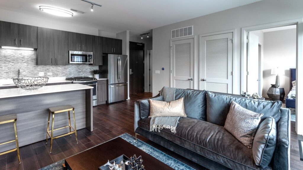 Modern open-concept living room and kitchen; includes a gray sofa, coffee table, kitchen island with stools, stainless steel appliances, and dark wood cabinetry.