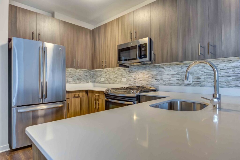 Modern kitchen with stainless steel appliances, wood cabinets, tiled backsplash, and a white countertop with a built-in sink.