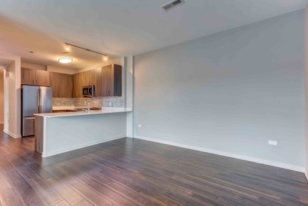 Empty modern kitchen and dining area with hardwood flooring, stainless steel appliances, gray cabinets, and a tiled backsplash. Walls are painted light gray.