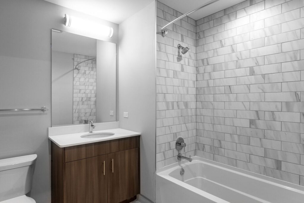 Modern bathroom with a shower-tub combo and gray tiled wall, a wooden vanity with a white countertop and sink, a large mirror, and a toilet.
