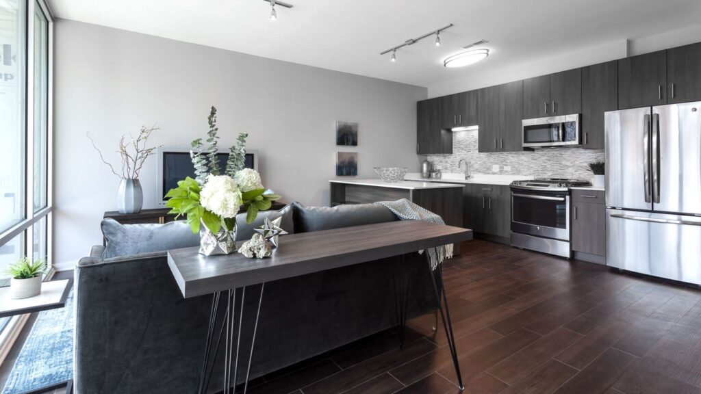 Modern open-concept living room and kitchen with dark wood floors, stainless steel appliances, gray sofa, and decorative plants on a console table.