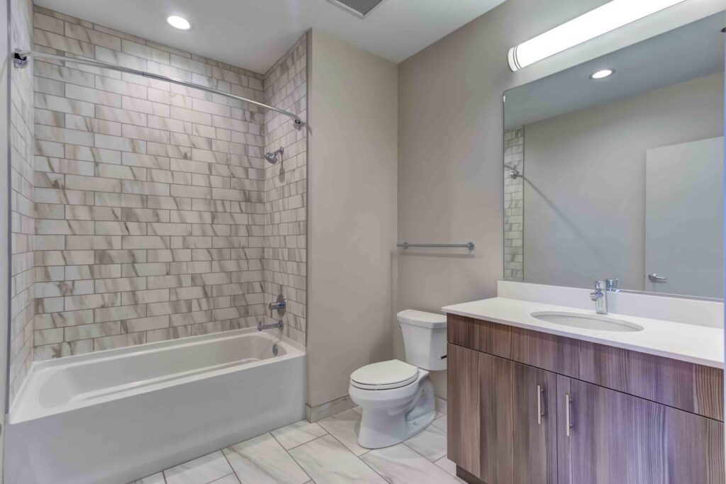 Modern bathroom with tiled shower-bathtub combo, toilet, and wooden vanity with single sink. Bright lighting and neutral color palette.
