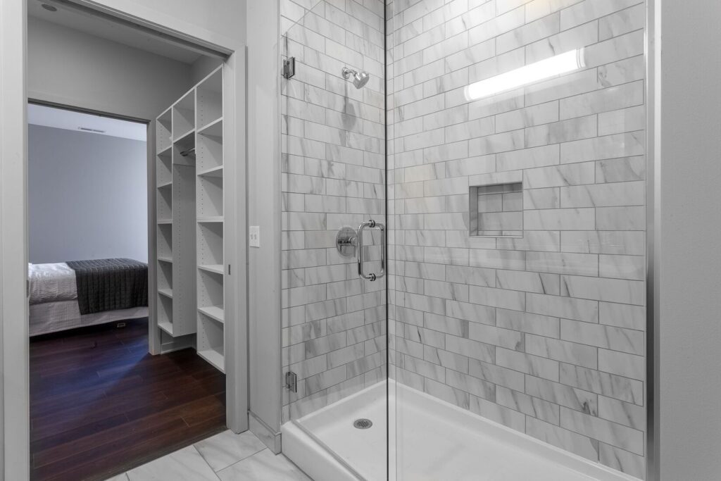 Modern bathroom with a glass-enclosed shower featuring light gray tile. In the background, there is a glimpse of a bedroom and open closet.