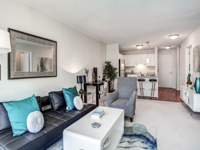 Modern living room with black sofa, turquoise pillows, armchair, white coffee table, and open kitchen in the background. Mirror, decor, and rug complete the space.