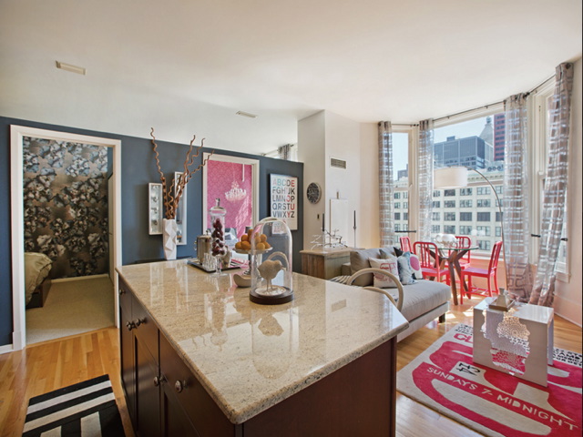 Modern apartment interior with an open-plan living area featuring a kitchen island, a sofa, and a dining space. Large windows provide city views. Bedroom visible in the left background.