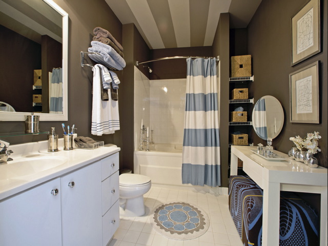 Modern bathroom with white fixtures, striped shower curtain, and brown walls. Shelving with storage baskets, wall mirror, and decorative accents. Towels are neatly stacked above the toilet.