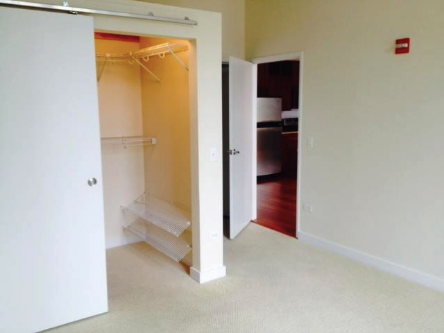 A room with an open closet featuring wire shelves and hangers, beige carpet, and a view into a kitchen with wooden flooring through an open door.