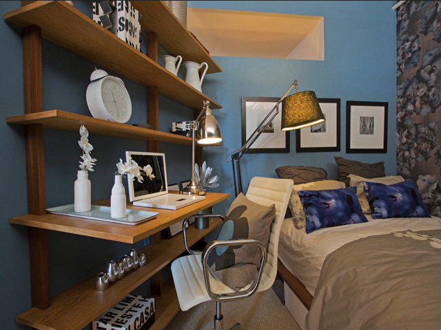 Cozy bedroom with blue walls features a bed, decorative pillows, wood shelves with books and decor, a desk with a laptop, and a floor lamp.