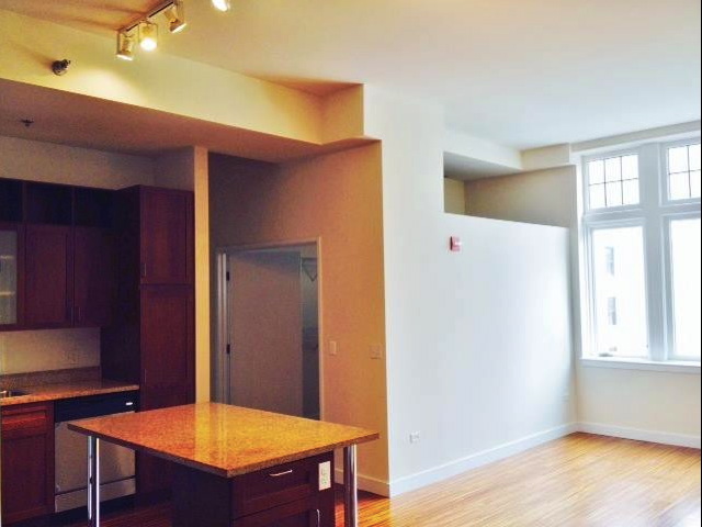 Modern apartment interior with kitchen featuring wooden cabinets and an island. Large windows on the right allow natural light to fill the open space, highlighting the wooden flooring.