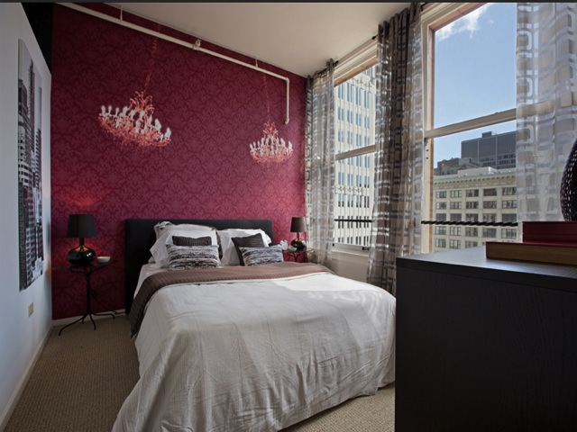 Stylish bedroom with a red accent wall, two chandeliers, a bed with white and brown bedding, two black side tables with lamps, and large windows with city views.