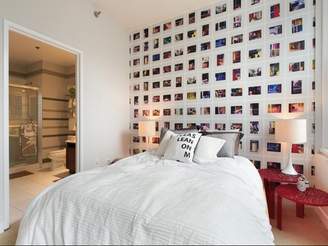 Modern bedroom with a white bed, a wall covered in colorful photos, a red nightstand with a lamp, and an open door leading to a bathroom. Pillow reads "PLEASE LEAN ON ME.
