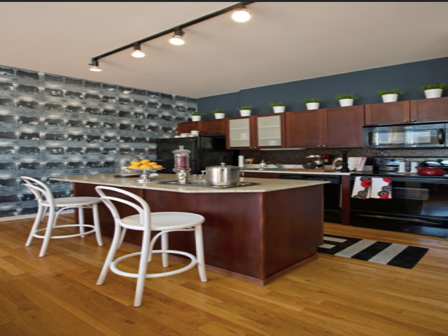 Modern kitchen with wooden flooring, island with white chairs, dark cabinets, and appliances. Potted plants on cabinets and a black-and-white patterned wall in the background.