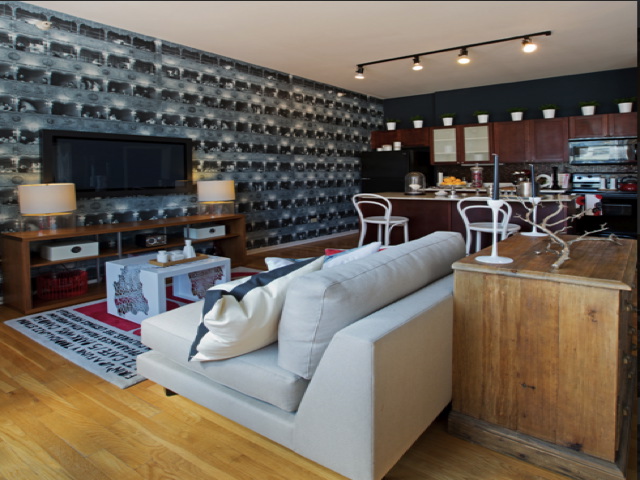 Modern living room with a white sofa, patterned wall, flat-screen TV, and kitchen area. Wooden flooring, white chairs, and a mix of contemporary and industrial decor elements are present.