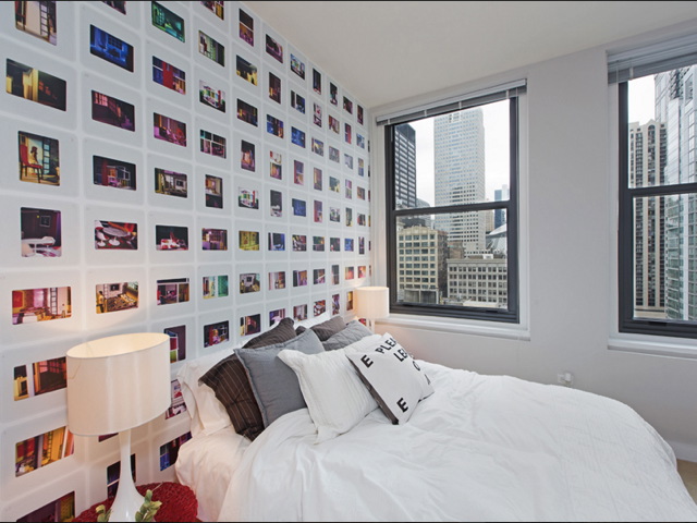 A modern bedroom with a city view, featuring a bed with white linens, two lamps, and a wall covered in photo prints, next to large windows showing skyscrapers.