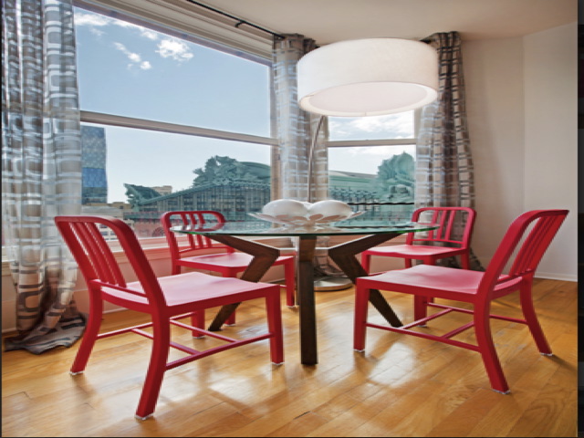 A round glass table with decorative items is surrounded by four red chairs on a wooden floor. Large windows with patterned curtains reveal an urban view. A modern ceiling light hangs above.
