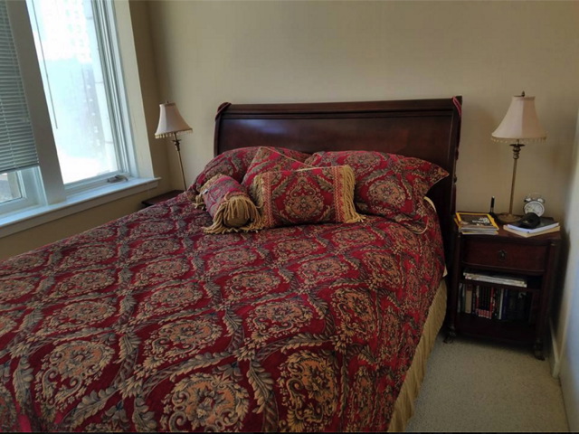 A bedroom with a wooden bed frame, patterned red and gold bedding, two lamps, and a nightstand with books and an alarm clock.