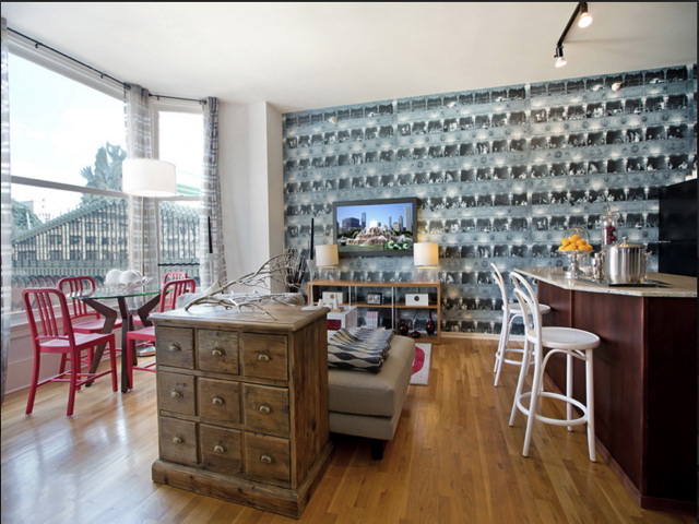 A living room with wooden flooring, a patterned wallpaper, and a small TV. Includes a dining table with red chairs, a wooden chest, and a bar area with white stools. Large windows on the left.