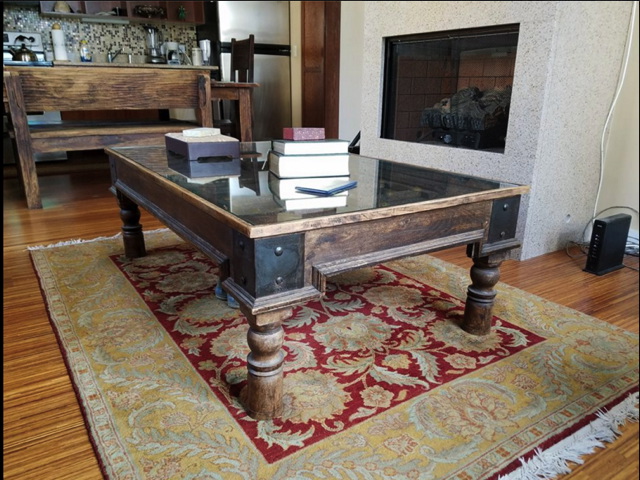 A wooden coffee table with a glass top is centered on an ornate rug. Several books and a box are on the table. A fireplace and dining area are visible in the background.