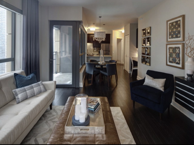 A modern living room with a white couch, blue armchair, and patterned rug. A dining table with blue chairs is in the background. Decor includes art on walls and a large mirror.