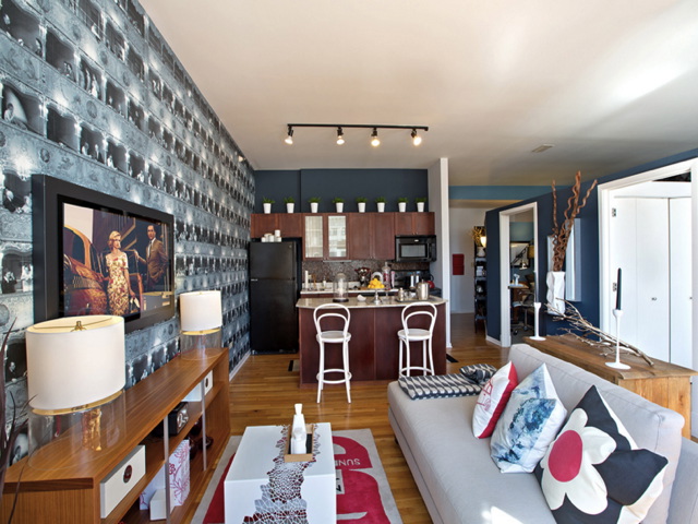 Modern living room with black and white patterned wall, colorful cushions on a gray sofa, a wall-mounted TV, kitchen with dark cabinetry, and barstools at a counter.