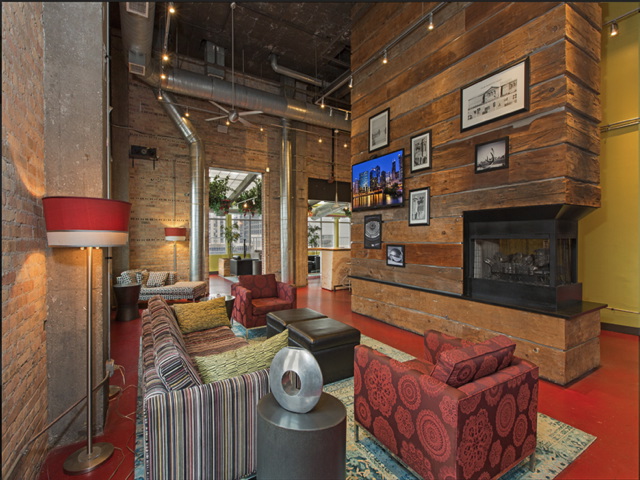 Stylish lounge with red flooring, patterned sofas, modern sculptures, and a TV. Exposed ductwork and brick walls create an industrial look. Framed photos decorate the wooden accent wall.