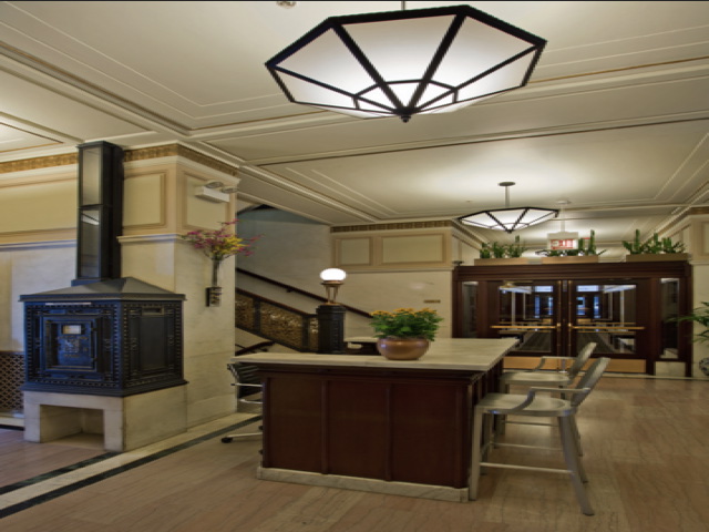 Building lobby with a large central wooden desk, bar stools, decorative lamp above, ornate heater on the left, and a staircase leading upstairs. Plants and lighting fixtures add ambiance.