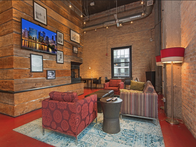 Modern rustic living room with exposed brick walls, red carpet, and eclectic furniture. A TV and framed pictures are mounted on a wooden wall. Large window and lamps provide lighting.