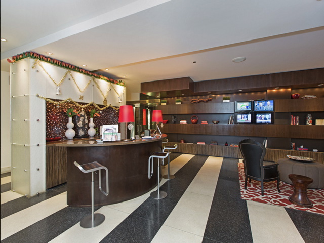 Modern hotel lobby with a curved reception desk adorned with vases and red lamps. Shelves on the right display books and decorative items. Black and white flooring with a red-patterned rug.