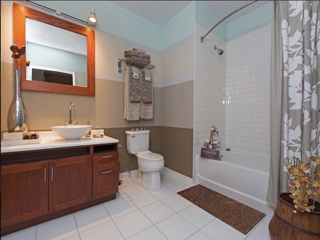 Bathroom with a wooden vanity, vessel sink, modern light fixture, toilet, tub-shower combo with floral curtain, and towels stacked on a wall shelf. White tile floor and beige wall.