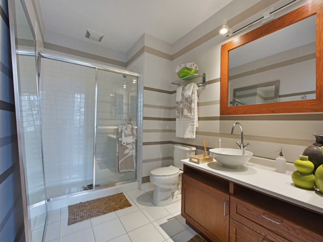 Modern bathroom with glass shower, wooden vanity with vessel sink, and large mirror. Striped walls, white tiled floor, and green decor accents.