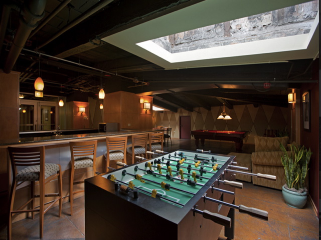 A recreation room with a foosball table, a bar with stools, a pool table, pendant lighting, and a skylight above.