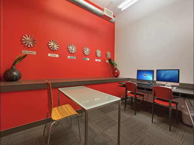 Modern office space with a red wall featuring decorative clocks labeled with city names. A table with a chair is in the foreground, and two computers are on a desk to the right.