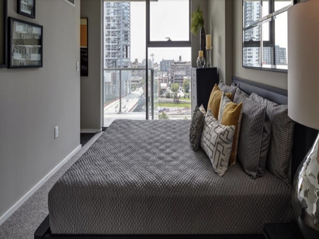 Minimalist bedroom with a large bed, decorative pillows, and cityscape view through a window. Modern decor with neutral tones and wall art.