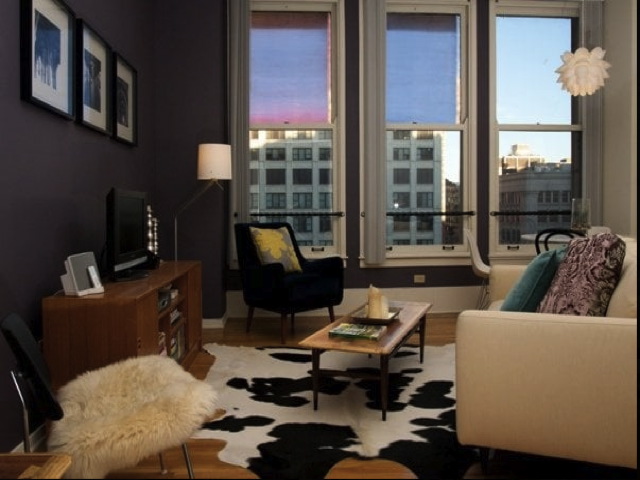 Cozy living room with a modern style, featuring a beige sofa, wooden coffee table, TV stand, and a black and white rug. Large windows with colorful blinds overlook a cityscape.