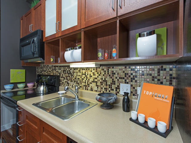 Modern kitchen with wooden cabinets, mosaic tile backsplash, stainless steel sink, electric stove, and microwave. A cookbook and measuring cups are on the counter.