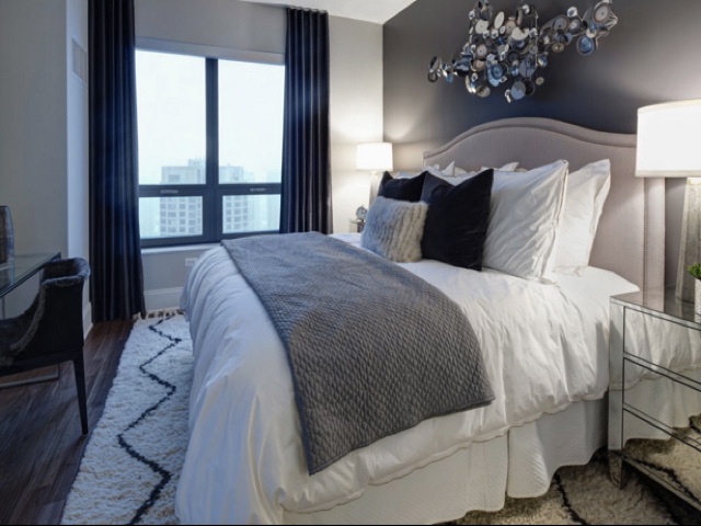 Modern bedroom with a large bed, dark pillows, and a gray blanket. Two lamps flank each side. Window with dark curtains shows a cityscape. Decorative wall art above the bed.