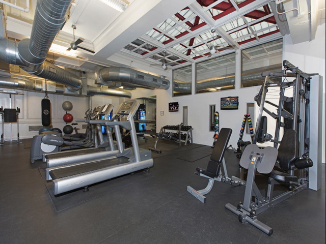 A gym with treadmills, weight machines, dumbbells, exercise balls, and a flat-screen TV in a room with exposed ductwork and a high ceiling.