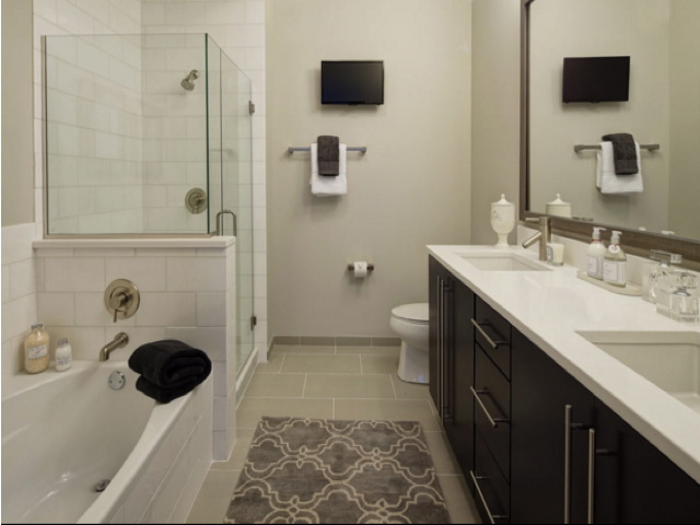 Modern bathroom with a glass shower, bathtub, dual-sink vanity, and wall-mounted TV. Neutral tones with a patterned rug and towel rack above the toilet.