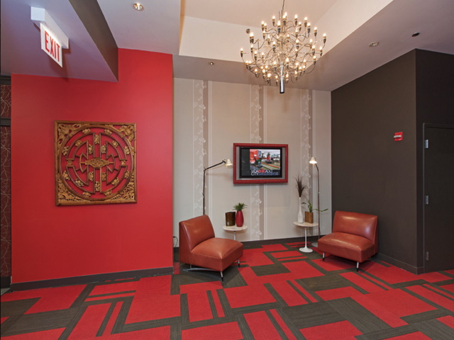 A modern lobby with a red and grey patterned floor, two brown chairs, a wall art piece, a wall-mounted lamp, and a chandelier. An exit sign is visible above a door.