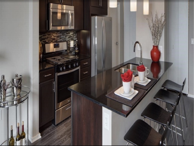 Modern kitchen with dark cabinets, stainless steel appliances, and a black countertop island. Two bar stools, decorative vases, and cups with red napkins on the island. Wine bottles on a side shelf.