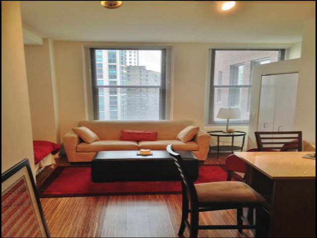 Small apartment with a beige sofa, red rug, and wooden floors. A bed is partially visible on the left. Two windows and a small table with chairs are present.