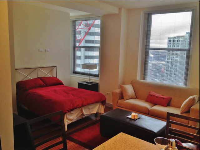 A small studio apartment with a bed, couch, and table. Large windows reveal a city view. A red comforter covers the bed, and a rug accents the floor.
