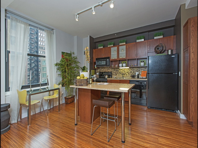 Modern kitchen with wooden cabinets, black appliances, and an island with seating. Dining table with two yellow chairs by the window. Potted plant and city view outside.