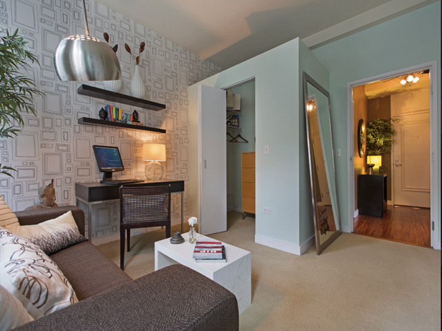 A modern living room with a patterned accent wall, desk with computer, large mirror, sofa, white coffee table, and open closet. Light green walls and ambient lighting create a cozy atmosphere.